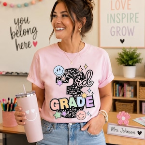May include: A light pink short-sleeve t-shirt featuring a graphic with the text 3rd GRADE, surrounded by colorful stars and smiley faces. The shirt is paired with blue denim jeans and a matching light pink tumbler with a white smiley face design.