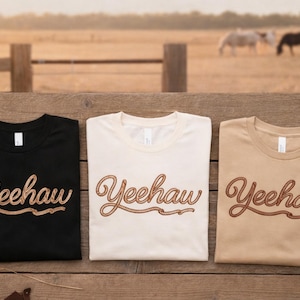 May include: Three t-shirts in black, cream, and tan, each with the word "Yeehaw" in a brown, rope-style font. The shirts are displayed on a wooden surface with a rural backdrop featuring horses.