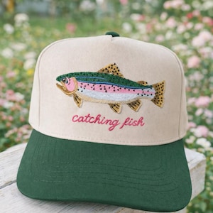 Könnte beinhalten: Beige und grüne Baseballkappe mit einem gestickten Regenbogenforellen-Design. Auf der Kappe steht in rosa Schriftzug "catching fish". Die Mütze liegt auf einer Holzoberfläche.