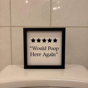 Peut inclure: Un panneau encadré avec une bordure noire et un fond blanc. Le panneau présente cinq étoiles noires au-dessus du texte "Would Poop Here Again" en noir, police sans-serif. Le panneau est placé sur des toilettes blanches.