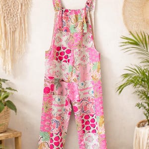 Peut inclure: Combinaison rose à motifs patchwork avec des motifs floraux et géométriques, suspendue à un cintre en bois. La combinaison a des bretelles réglables et des jambes larges. Le tissu est rose, blanc et vert.