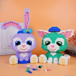 May include: Two assembled bunny figures made of small building blocks. One is purple with pink accents, the other is green with pink accents. Both have large ears, blue bows, and white paws. Scattered blocks are in front.