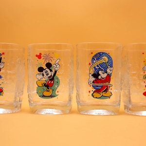 May include: Four clear drinking glasses with colorful Mickey Mouse designs. Each glass features a different pose of Mickey Mouse, with vibrant red, yellow, blue, and black colors. The glasses have a textured appearance and are set against a solid orange background.