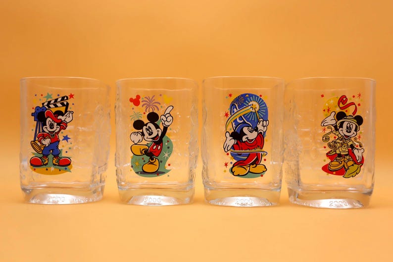 May include: Four clear drinking glasses with colorful Mickey Mouse designs. Each glass features a different pose of Mickey Mouse, with vibrant red, yellow, blue, and black colors. The glasses have a textured appearance and are set against a solid orange background.