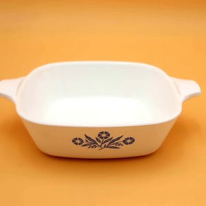 May include: A white square casserole dish with two handles. The dish has a floral design in dark blue on the front. The background is a solid orange color.