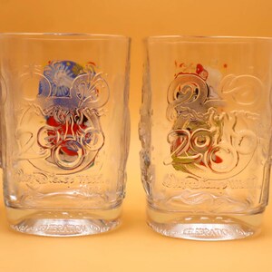 May include: Clear glass tumblers with embossed designs. Each glass features a colorful graphic with the number "20" and the words "Disney World" and "Celebration". The glasses have a slightly textured surface.