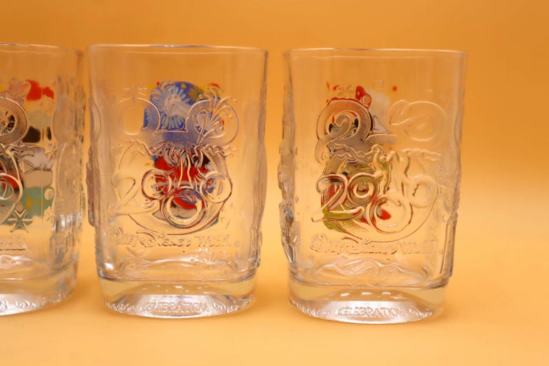 May include: Clear glass tumblers with embossed designs. Each glass features a colorful graphic with the number "20" and the words "Disney World" and "Celebration". The glasses have a slightly textured surface.