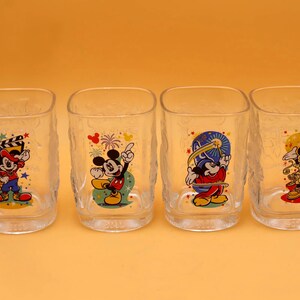 May include: Four clear, square-shaped drinking glasses with textured sides. Each glass features a different colorful image of Mickey Mouse in various poses. The glasses are arranged in a row against a solid orange background.