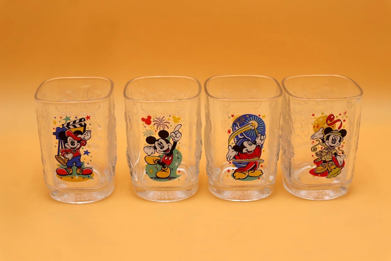 May include: Four clear, square-shaped drinking glasses with textured sides. Each glass features a different colorful image of Mickey Mouse in various poses. The glasses are arranged in a row against a solid orange background.