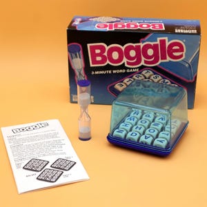 May include: Boggle word game set. The box is black and blue with the word "Boggle" in pink. Includes a clear plastic game grid, dice with letters, a 3-minute timer, and instructions.