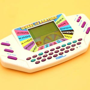 May include: A white handheld electronic Wheel of Fortune game by Tiger with a central LCD screen, a QWERTY keyboard featuring purple buttons, and additional purple function buttons on the sides. The device displays the Wheel of Fortune logo.