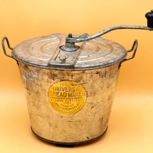 May include: Vintage Universal Bread Maker, a metal bread-making machine with a hand crank and a black handle. The bread maker has a round, aged appearance with a faded yellow label that reads "Universal Bread Maker". The machine has two handles.