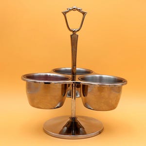 May include: A silver-colored condiment server with three round bowls and a handle. The bowls are arranged around a central support and sit on a circular base. The handle has a decorative design.