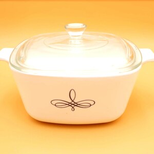 May include: A white casserole dish with a clear glass lid and a decorative black swirl design. The dish has two handles and is on a yellow background. This vintage kitchenware is perfect for baking and serving.