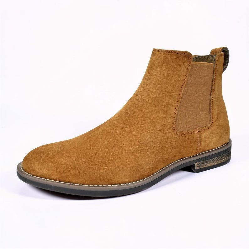 Peut inclure: Une botte Chelsea unique en daim de couleur beige. La botte pr&eacute;sente un bout arrondi, un panneau lat&eacute;ral &eacute;lastique et une tirette &agrave; l'arri&egrave;re. La semelle est fonc&eacute;e avec un d&eacute;tail de couture de couleur claire. La botte est sur fond blanc.