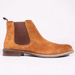 Peut inclure: Une botte Chelsea en daim marron clair avec une languette et un panneau latéral élastique. La botte a un bout arrondi et un petit talon empilé. La semelle est de couleur claire, et la botte est sur fond blanc.