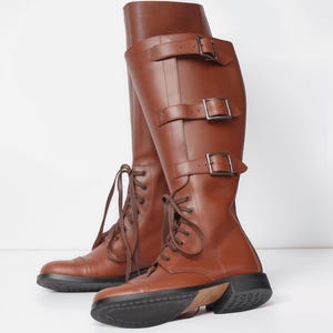 Peut inclure: Une paire de bottes hautes en cuir marron. Elles sont dotées d'un laçage à l'avant, de trois sangles à boucle et d'une semelle épaisse noire. Les bottes ont un talon compensé et semblent neuves. La hauteur des bottes est d'environ 40 cm.
