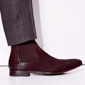 Könnte beinhalten: Burgunderfarbene Chelsea-Stiefel aus Wildleder mit einem Lederbesatz mit Krokodiltextur an der Ferse. Der Stiefel hat eine elegante, spitze Zehenpartie und einen niedrigen Absatz. Die Stiefel werden mit grauen Hosen kombiniert.