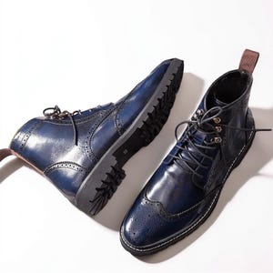 May include: Two blue leather boots with brogue detailing and a lace-up design. The boots have a dark sole and a pull tab at the back. The boots are displayed on a white background.
