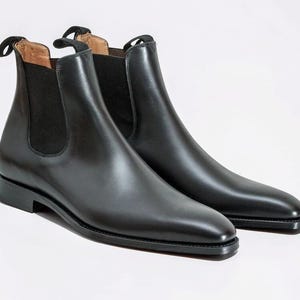 Könnte beinhalten: Ein Paar schwarze Chelsea-Stiefel aus Leder. Die Stiefel haben eine glatte, polierte Oberfläche, elastische Seiteneinsätze und Zuglaschen hinten. Die Sohlen sind dunkel, und die Stiefel stehen vor weißem Hintergrund.