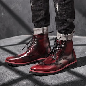 May include: A pair of burgundy leather boots with black laces. The boots have a wingtip design and a black sole with a red trim. The boots are worn with dark denim jeans.