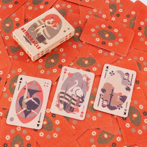 Puede incluir: Una baraja de cartas Retroracle con una caja beige y dorsos rojos. Las cartas presentan ilustraciones de figuras y símbolos. La caja dice "Retroracle Limited Edition". Las cartas están esparcidas sobre una superficie roja con patrones florales.