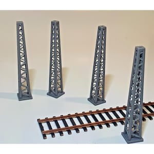 May include: Four grey miniature model train towers with a lattice design are arranged near a section of brown and black model train tracks. The towers are of varying heights and angles, suggesting a diorama or model railway scene.