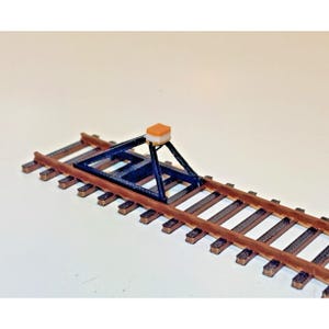 May include: A miniature model train track with brown wooden ties and metal rails. A dark blue metal structure with an orange square on top sits on the track. The model is set against a white background.