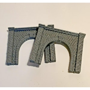 Könnte beinhalten: Zwei graue Miniatur-Steinbogen, wahrscheinlich für eine Modelleisenbahn oder ein Diorama. Die Bögen haben ein strukturiertes, verwittertes Aussehen, mit einem rechteckigen Schlussstein über dem Bogen. Die Bögen bestehen aus einem steinähnlichen Material.