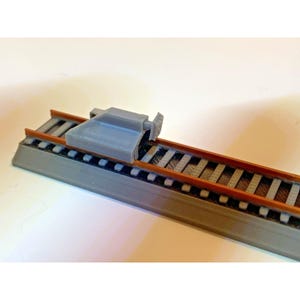 May include: A gray 3D-printed model of a railroad track section with a buffer stop. The track features brown rails and gray ties. The buffer stop is a gray, rectangular structure designed to halt trains.
