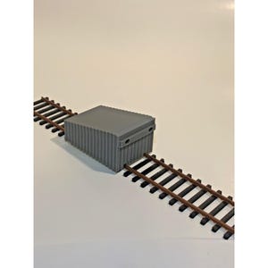 May include: A gray, rectangular model shipping container with vertical ridges, positioned between two sections of miniature train tracks. The tracks are brown and black, and the container has two small holes on its side.