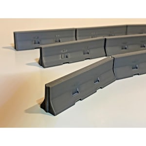May include: A collection of gray plastic road barriers. Each barrier has a rectangular shape with a flat top and a slightly angled front. The barriers are designed to interlock, creating a continuous line.