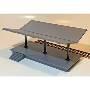 May include: A gray model train station platform with a sloped roof supported by black pillars. The platform has a small set of stairs and is next to a section of brown model train tracks.