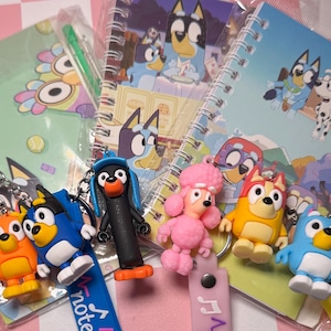 Puede incluir: Una colección de productos de Bluey, que incluye llaveros y cuadernos de espiral. Los llaveros presentan personajes de dibujos animados en varios colores. Los cuadernos tienen ilustraciones de dibujos animados en las portadas. Los artículos están colocados sobre una superficie rosa y blanca.