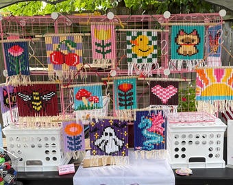 Puede incluir: Una colección de pequeños tapices rectangulares de cuentas exhibidos en una rejilla de alambre rosa. Cada pieza presenta diseños pixelados que incluyen cerezas, un pato amarillo, un perro, una abeja, setas, un corazón, un fantasma y un caballito de mar, con flecos.