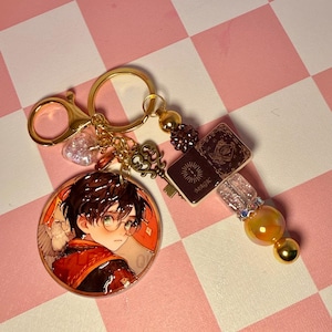 May include: A gold-colored keychain featuring a circular charm with a cartoon illustration of a character with glasses and a red and black outfit. The keychain includes a key charm, a heart-shaped charm, and charms with the word "MAGIC" on them.
