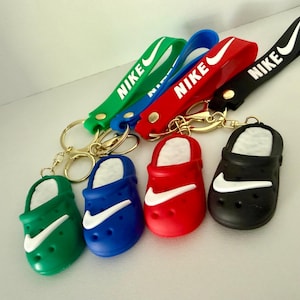 Può includere: Portachiavi con ciondoli a forma di scarpe in miniatura verdi, blu, rosse e nere. Ogni ciondolo ha un logo Nike bianco e un cinturino da polso abbinato con il logo Nike. Portachiavi color oro.