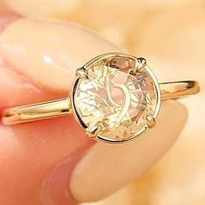 May include: A gold ring with a large, round, light-yellow gemstone. The gemstone is set in a four-prong setting and has a detailed, internal design. The ring band is thin and polished, and the overall design is elegant.