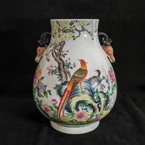 Puede incluir: Un jarrón de cerámica blanca grande con una pintura detallada y colorida de pájaros, flores y un árbol. El jarrón tiene asas con forma de cabeza de ciervo y una base ancha. La obra de arte presenta tonos vibrantes de naranja, verde, rosa y azul.