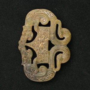 Può includere: Un ciondolo in giada scolpita con un design traforato a spirale. Il ciondolo è di colore verde pallido con motivi e dettagli intricati. La forma è asimmetrica con bordi arrotondati e un design centrale.