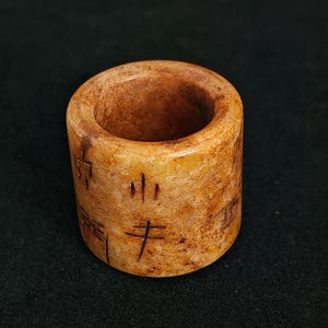 May include: A cylindrical, light brown stone ring with etched characters. The ring has a smooth, polished surface and a hollow center. The stone has variations in color, with darker brown areas. The ring is likely an antique or vintage item.