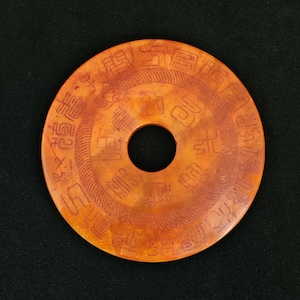 May include: A circular, orange-brown jade artifact with a central hole and intricate carvings. The surface features a series of symbols and patterns, likely of historical or cultural significance. The object is set against a dark background.