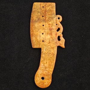 May include: A carved, antique jade artifact with a light brown and yellow color. The object has a handle and a decorative cutout design. There are several small holes and what appears to be engraved text on the surface. The artifact is set against a black background.