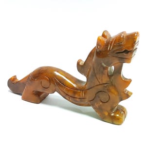 May include: An intricately carved, amber-hued sculpture of a mythical beast, possibly a dragon or lion. The figurine features detailed carving, with a stylized mane, open mouth, and curved form. The sculpture is set against a white backdrop.