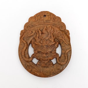 May include: A brown, oval-shaped pendant with a carved dragon design. The pendant features a detailed dragon head with flames and intricate patterns. It has a hole at the top for hanging and is made of a stone-like material.