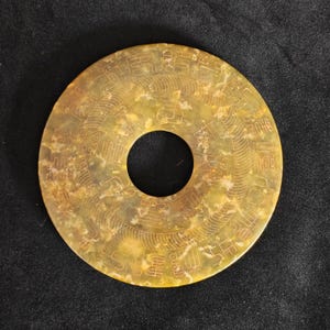 May include: A circular, antique jade artifact with a central hole. The jade is a mottled yellow-green color, with intricate carvings of Chinese characters and patterns across its surface. The artifact is set against a dark background.