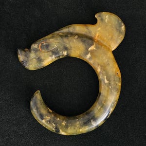 Peut inclure: Un pendentif en jade sculpté en forme de dragon stylisé. Le pendentif présente un mélange de teintes jaunes et vert foncé, avec un corps incurvé et une tête de dragon détaillée. La pièce est posée sur un fond noir.