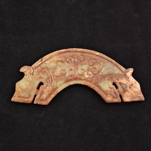 May include: A carved, arch-shaped jade artifact with a light green and brown color palette. The jade features intricate floral and scrollwork designs. The piece has two rectangular cutouts and two stylized animal head-like protrusions on each end.