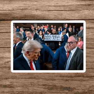 May include: A group of people in suits and dresses are gathered in a room. A person is holding a sign that reads "BLACK PEOPLE AREN'T APES!". Former President Donald Trump is in the foreground.