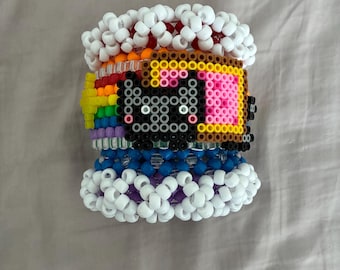 May include: A wide cuff bracelet constructed from colorful plastic pony beads. The design features a central black cat face icon, a rainbow-colored stripe pattern, and thick borders of white beads at the top and bottom edges of the cuff.